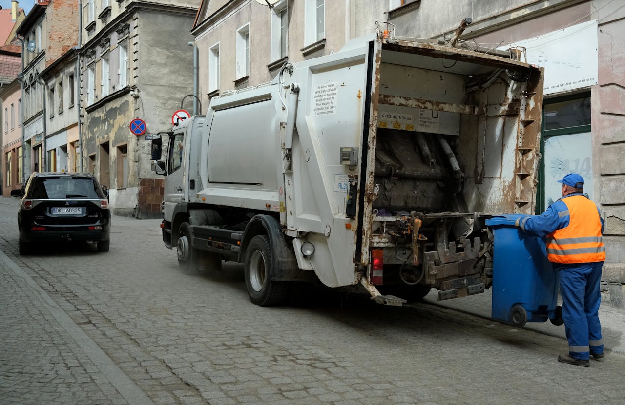 City street scene with garbage truck and worker handling waste bin.
