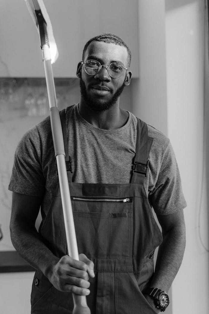 Black and white image of a bearded man in overalls holding a mop, conveying professionalism.
