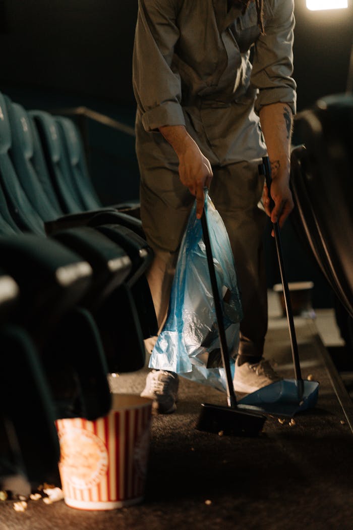 A worker cleans up spilled popcorn in a movie theater's aisle with a broom and dustpan.